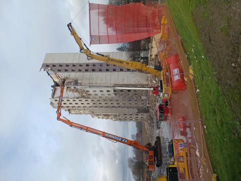 A colour photo of the towers during demolition. In the foreground, there is industrial fencing and a red net being lifted to control debris, with demolition in progress on the tower in the background. Only one of the towers is in frame, and over half of it is rubble.